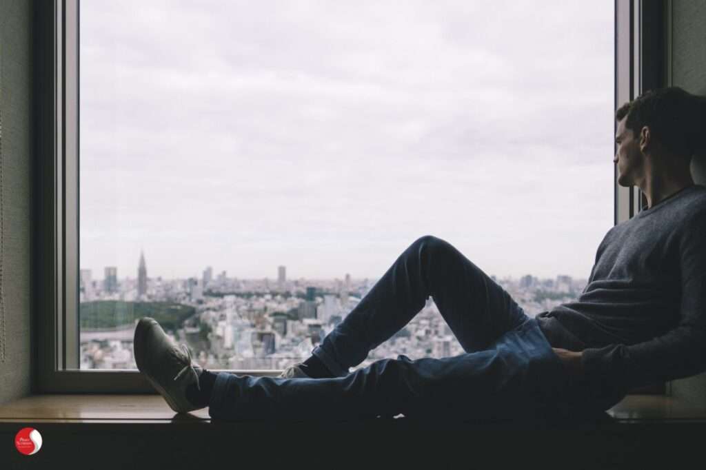 Homem sentado em silêncio observando a cidade pela janela, representando reflexão interior — como a psicoterapia pode te ajudar a ampliar a consciência emocional.