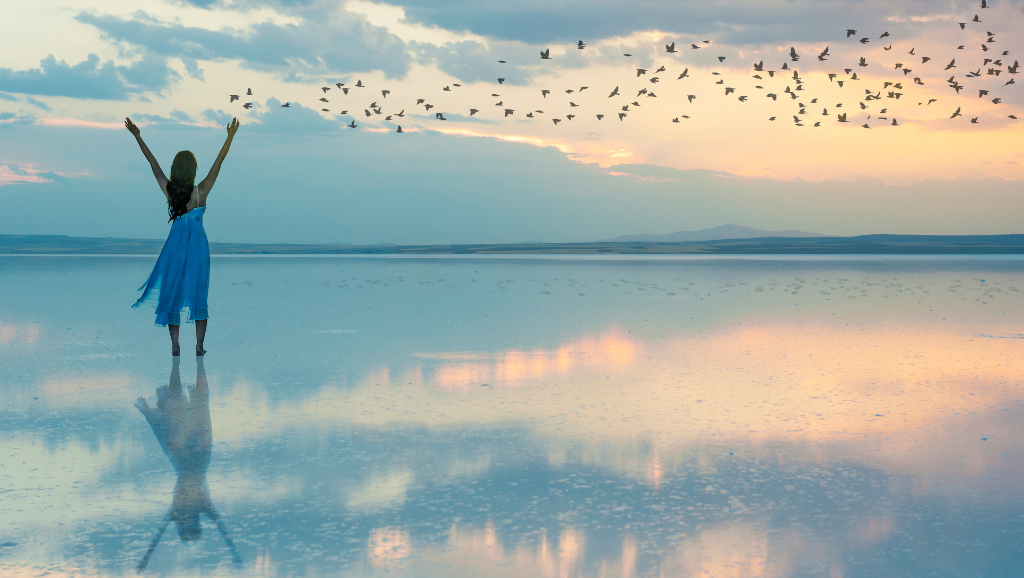 Mulher de braços abertos em lago reflexivo, pássaros voando, paz após superar o transtorno do pânico