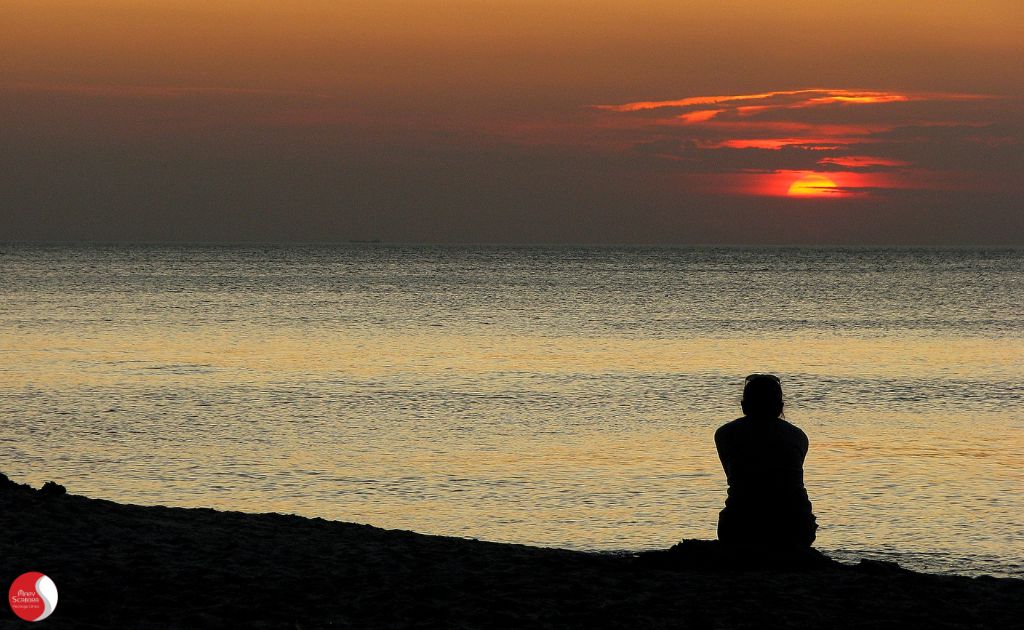 Pessoa sentada isolada à beira-mar ao pôr do sol. O aconselhamento psicológico para luto, perdas e transições de vida pode ajudar a encontrar suporte