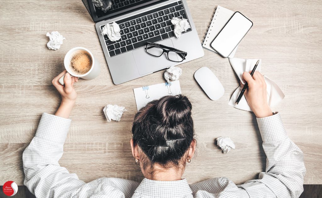 Pessoa exausta com a cabeça deitada na mesa, cercada por papel amassado, laptop e café, representando cansaço extremo e fadiga mental no trabalho.