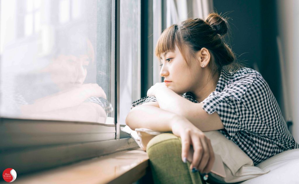 uma mulher olhando pela janela com expressão pensativa, simbolizando o isolamento emocional, a desconexão de familiares e amigos, e a solidão interna