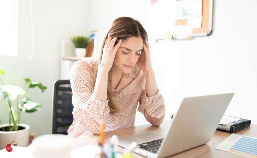 Mulher em ambiente de trabalho segurando a cabeça, demonstrando sintomas físicos do estresse, como dores de cabeça e fadiga, comuns em rotinas profissionais intensas.