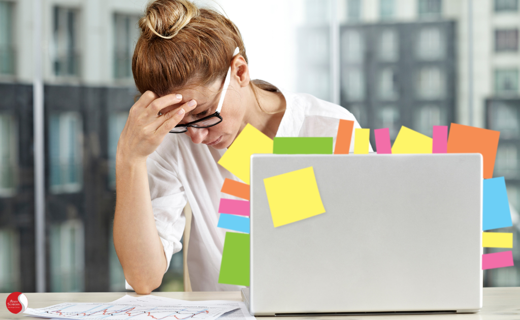 Mulher sobrecarregada em frente ao laptop com notas adesivas, representando os efeitos da busca pela perfeição desadaptativo, como estresse e exaustão.