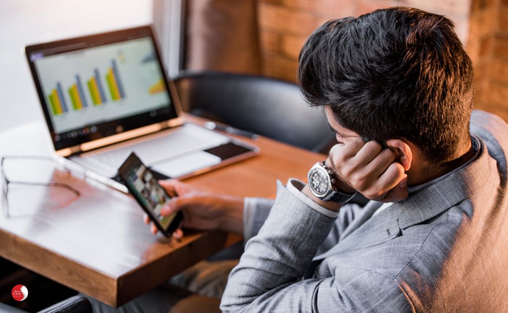 Homem procrastinando no trabalho ao olhar o celular, representando um dos sintomas do estresse, como dificuldade de foco e produtividade em tarefas importantes.