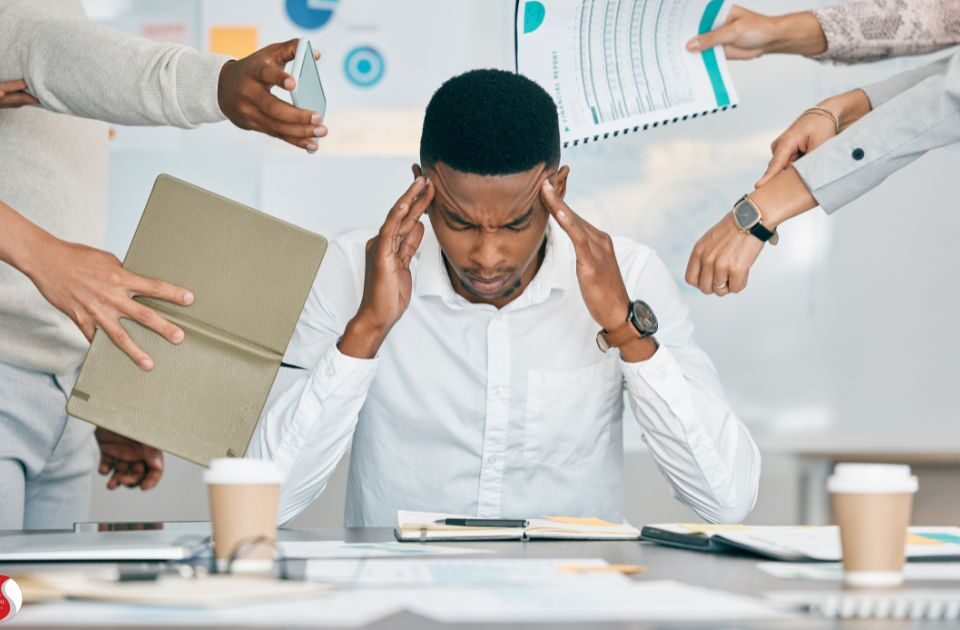 Homem pressiona as têmporas com expressão de estresse enquanto várias mãos ao redor apresentam tarefas e dispositivos, simbolizando a sobrecarga e pressão no ambiente de trabalho