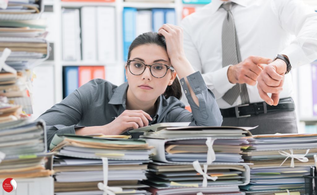 Mulher exausta cercada por pilhas de documentos enquanto colegas apontam para o relógio, representando a sobrecarga de trabalho e a pressão por produtividade.