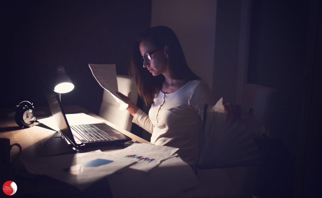 Mulher trabalhando tarde da noite, cercada por papéis e documentos, simbolizando a negligência do autocuidado e o comprometimento exagerado com o trabalho.