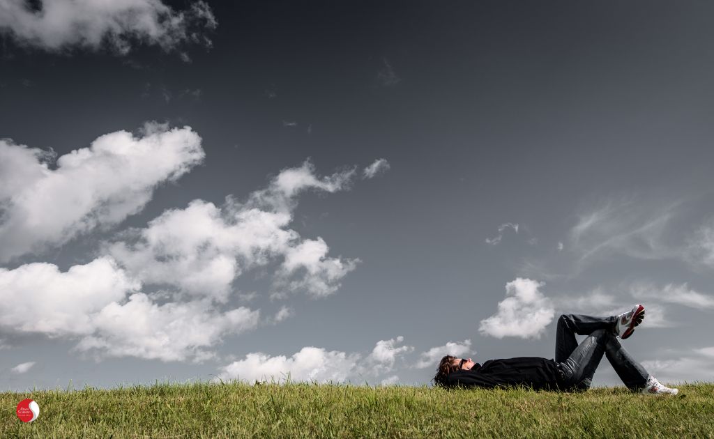 Pessoa deitada na grama sob um céu nublado, desfrutando de um momento de relaxamento ao ar livre, simbolizando a importância de pausas para o bem-estar e o equilíbrio emocional.